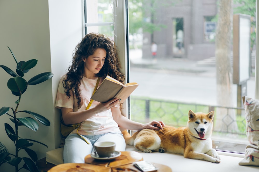 uma mulher sentada no parapeito de uma janela lendo um livro ao lado de dois cachorros