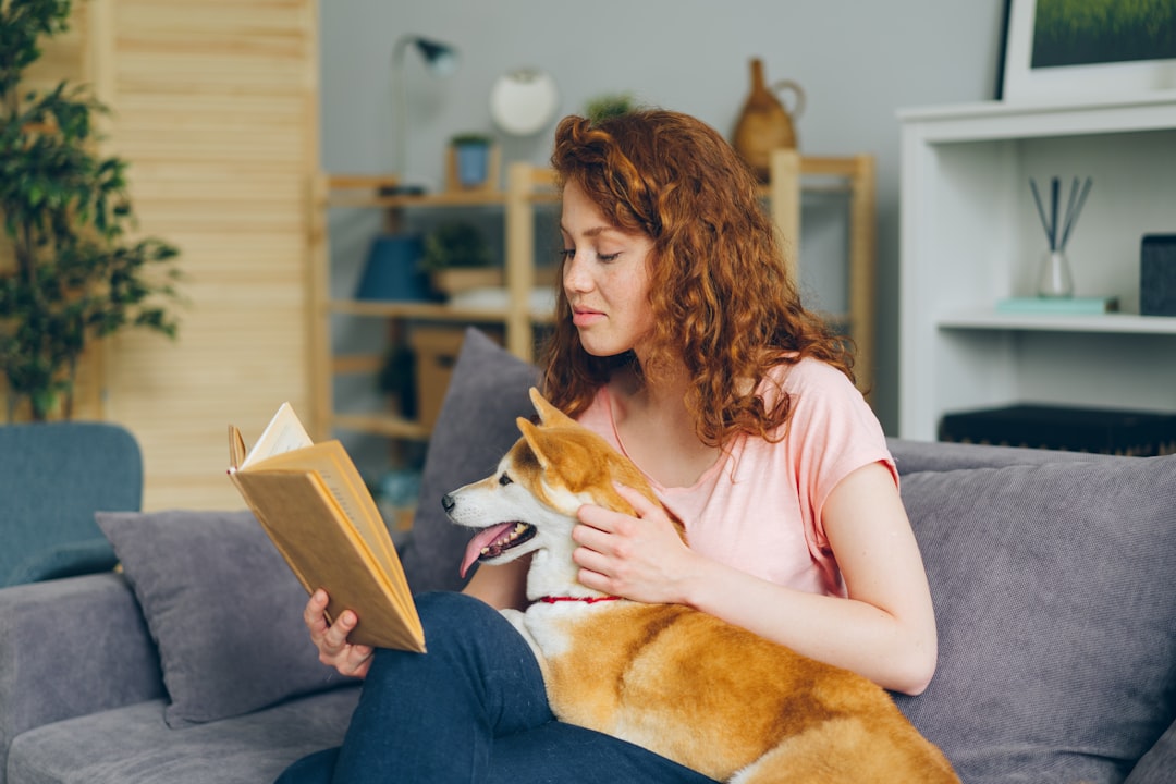 a woman sitting on a couch reading a book with her dog, Pretty woman reading book smiling stroking shiba inu puppy on so