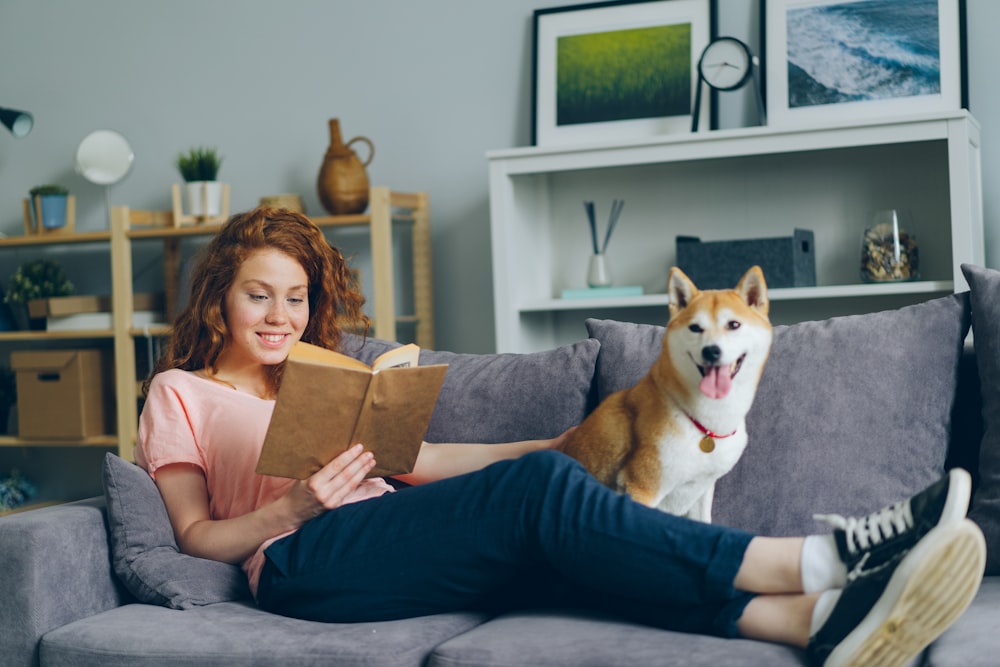 a woman sitting on a couch reading a book next to a dog