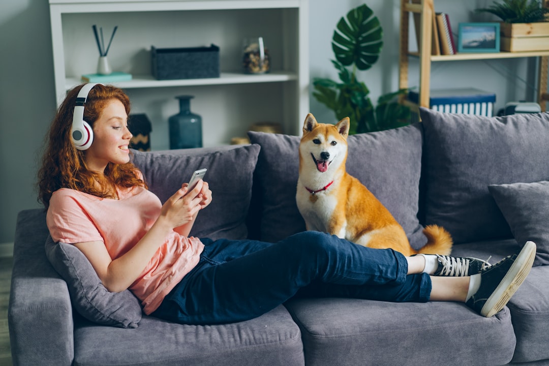 a woman sitting on a couch next to a dog, Student in headphones listening to music using smartphone on couch with dog