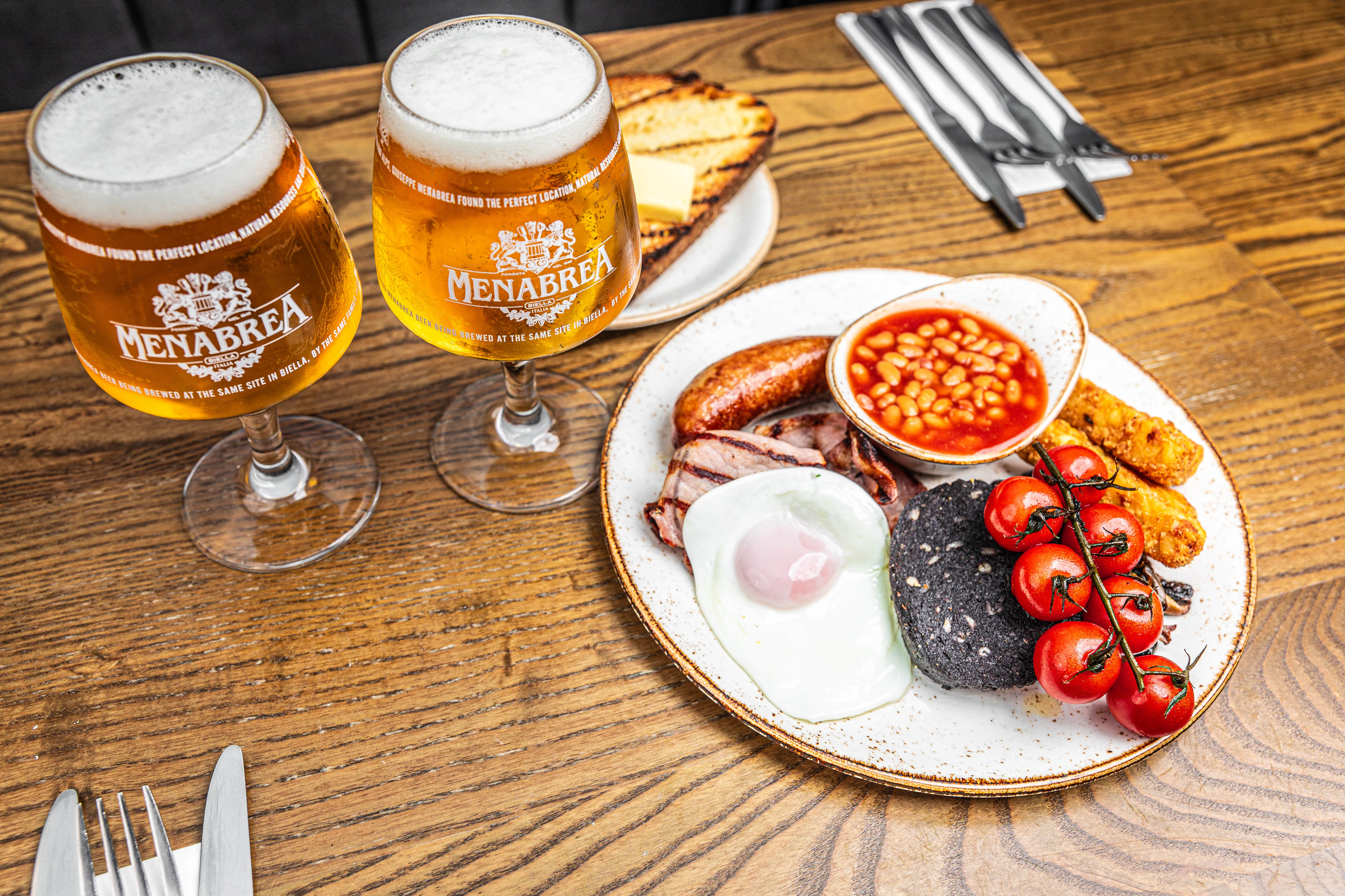 A full British fried breakfast on a pub table — sausage, bacon, fried egg, black pudding, baked beans, vine tomatoes and hash brown — served with two glasses of lager