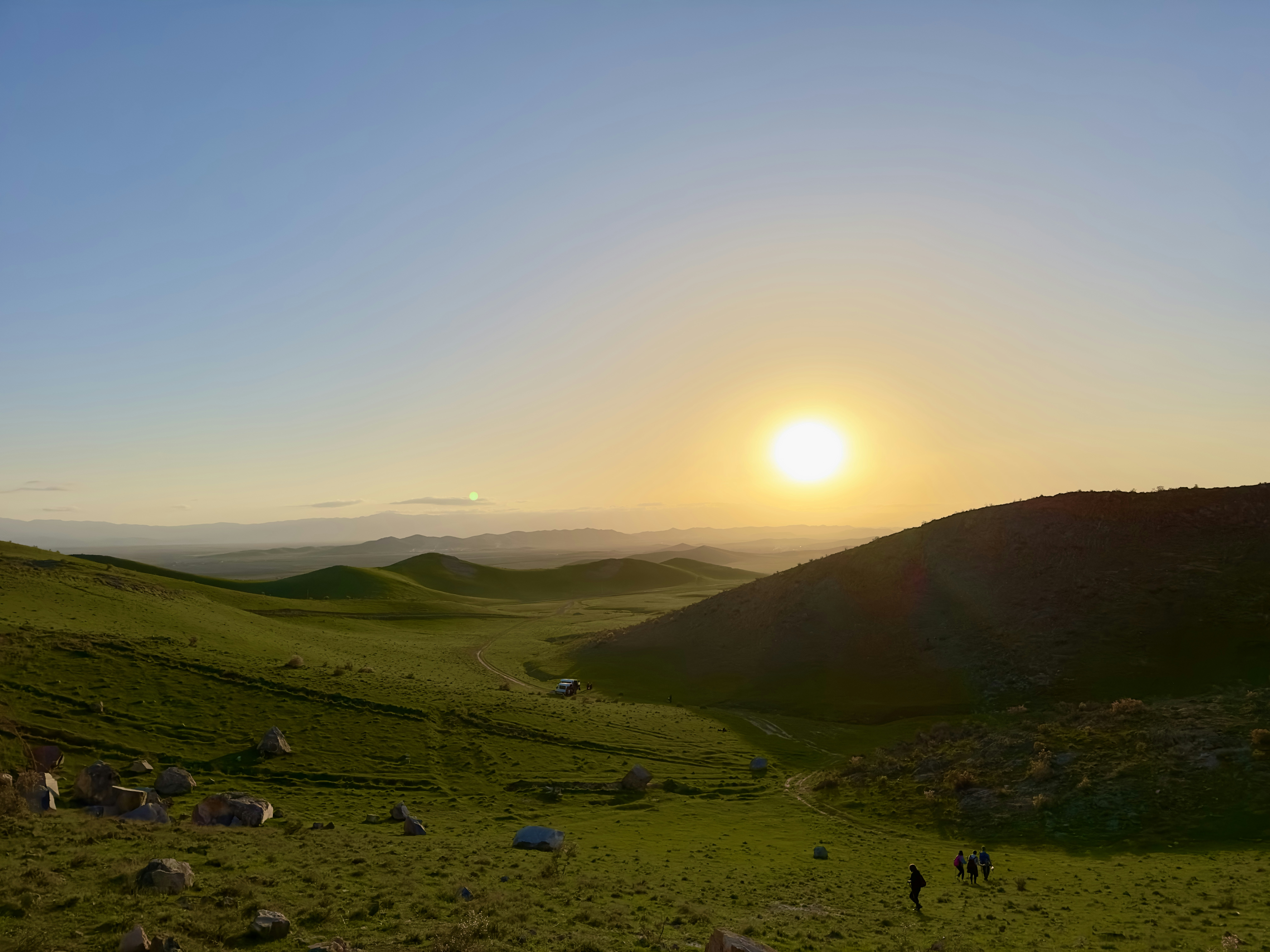 the sun is setting over a grassy hill, The setting sun bathes the hills of Aidarkul in Jizzakh in soft orange and pink hues, creating a captivating spectacle of contrast and harmony. The dark silhouettes of the hills are outlined against the bright blue sky, inviting a moment of pause to breathe in the magic of the moment. This sunset reminds us of the beauty of nature that can stop time, offering a sense of calm and serenity, immersing in reflections on the majesty of the world around us.