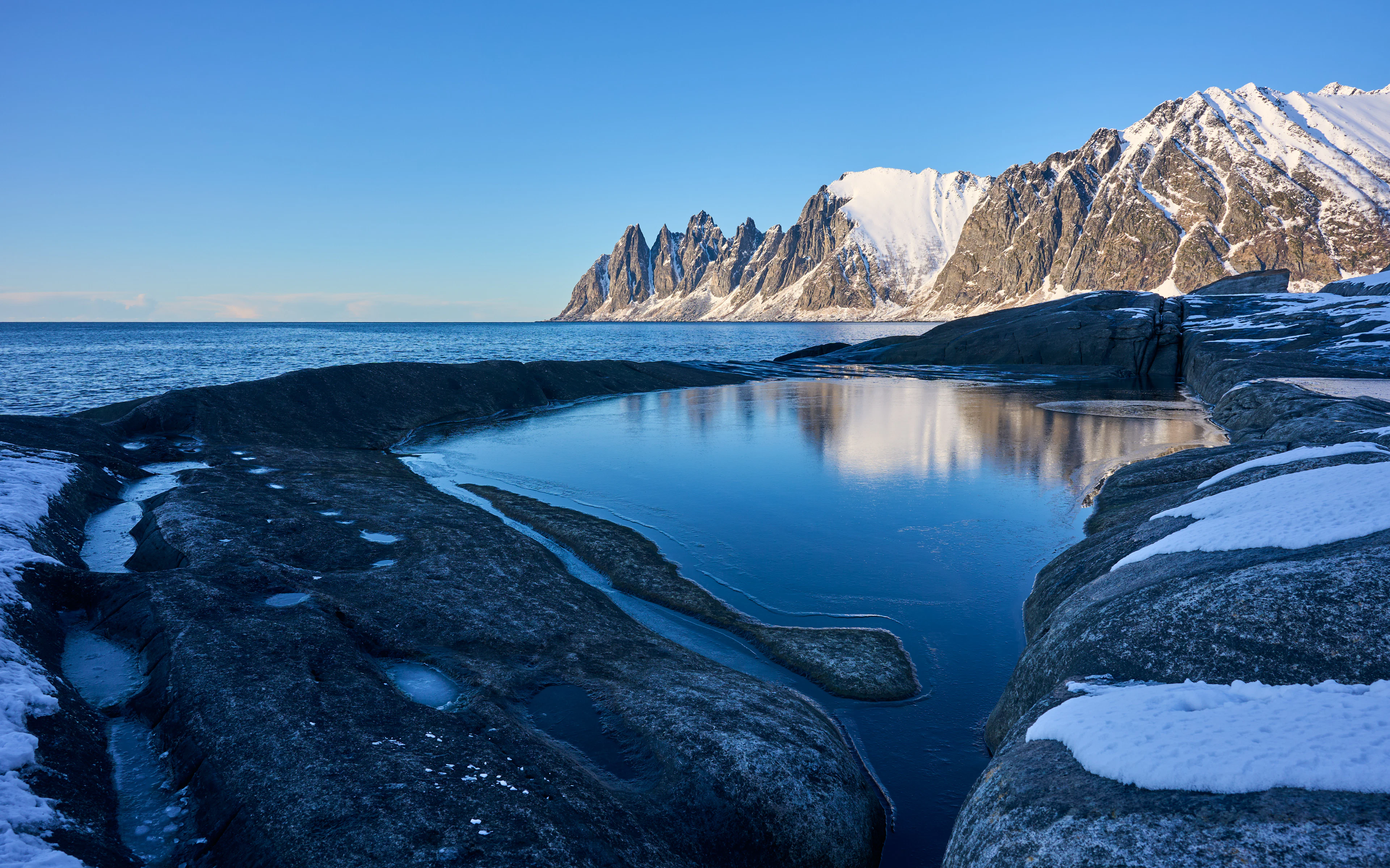 Jagged snow-covered peaks above ice-smoothed coastal rock and still water on Senja in winter
