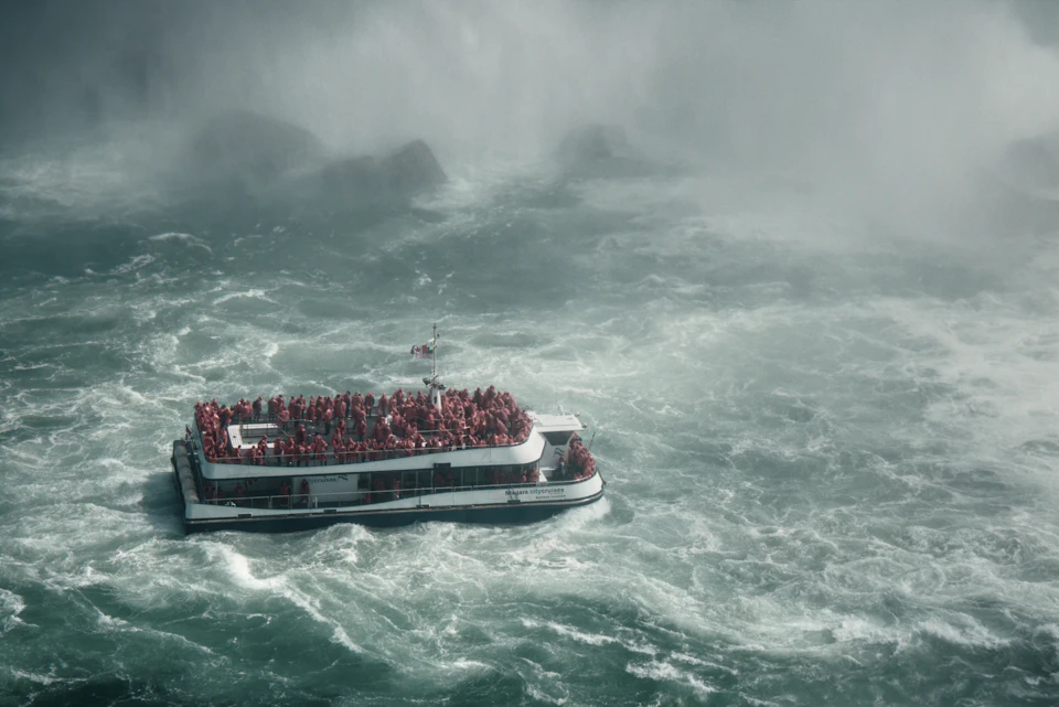 The image depicts a large white boat with multiple levels, filled with people wearing life jackets and standing on the deck. The boat is moving through rough waters, creating a sense of motion and adventure. In the background, there are rocks visible, adding to the ruggedness of the scene.