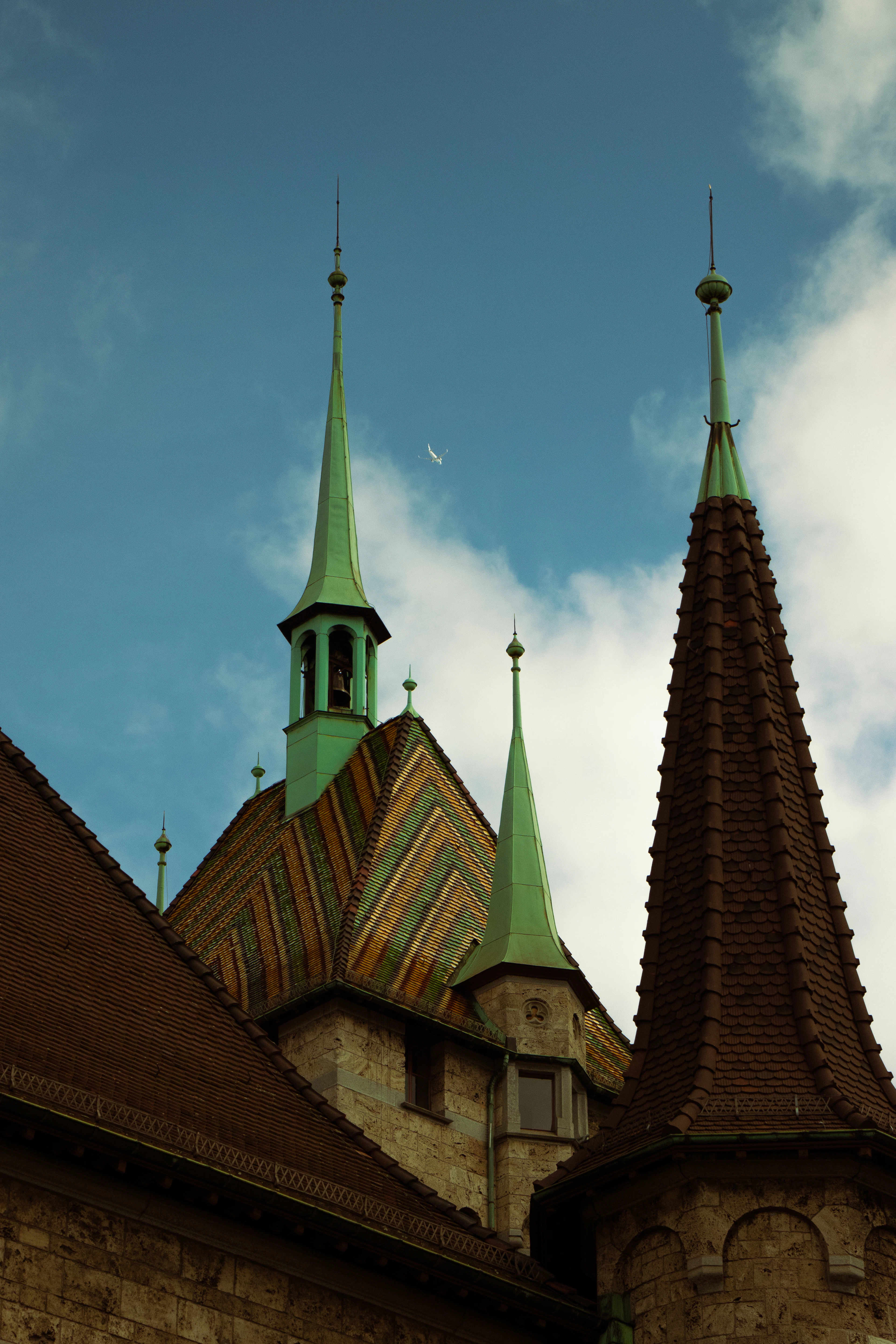 Green copper spires and a patterned tile roof rise against a blue sky as a bird flies past.