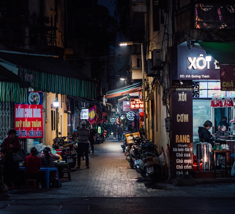 a narrow street at night with people sitting at tables