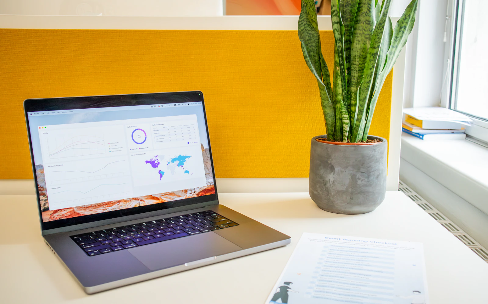 Laptop on a bright desk displaying an analytics dashboard beside a plant and printed checklist
