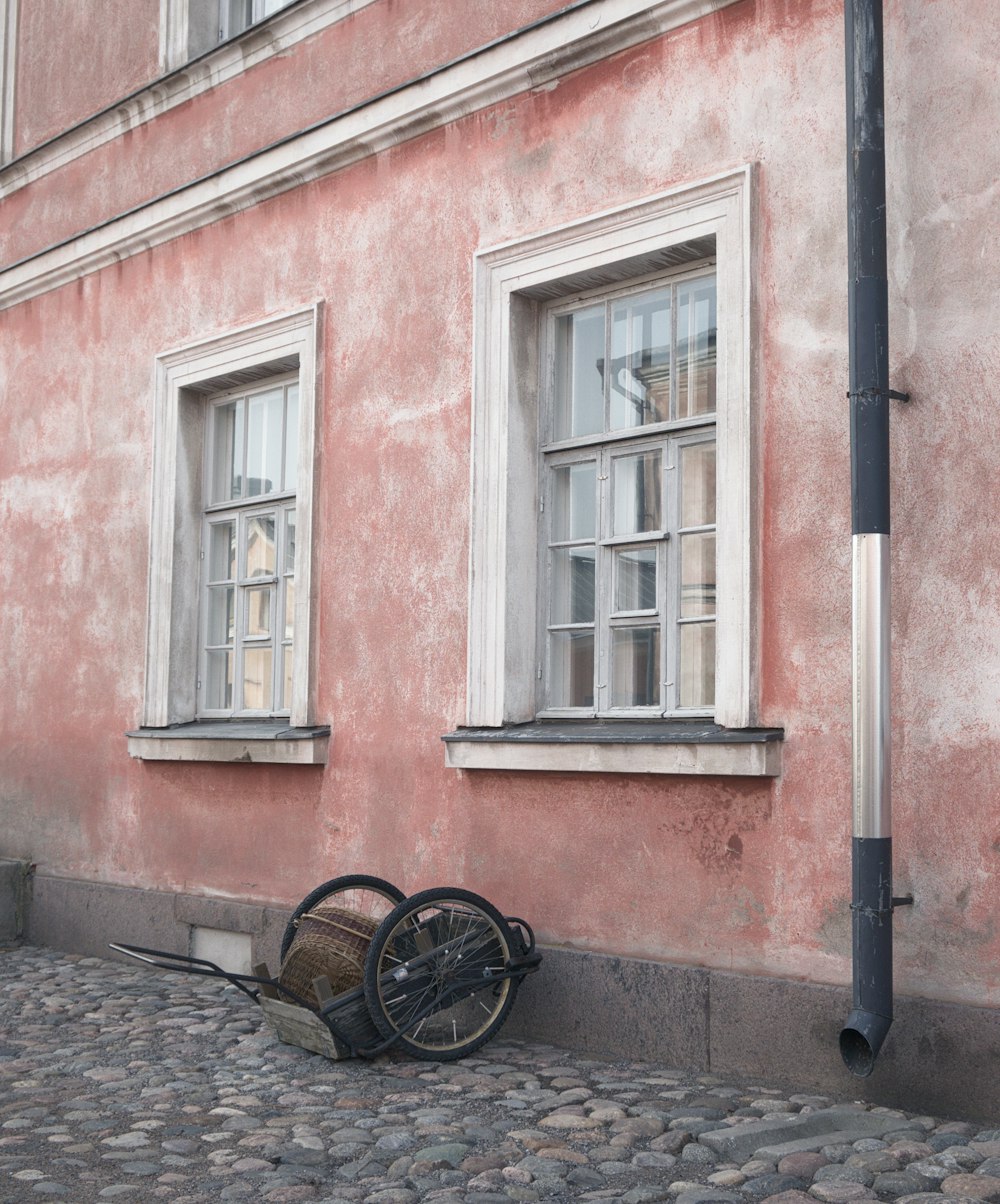 a wheelbarrow leaning against a pink building on a cobblestone street