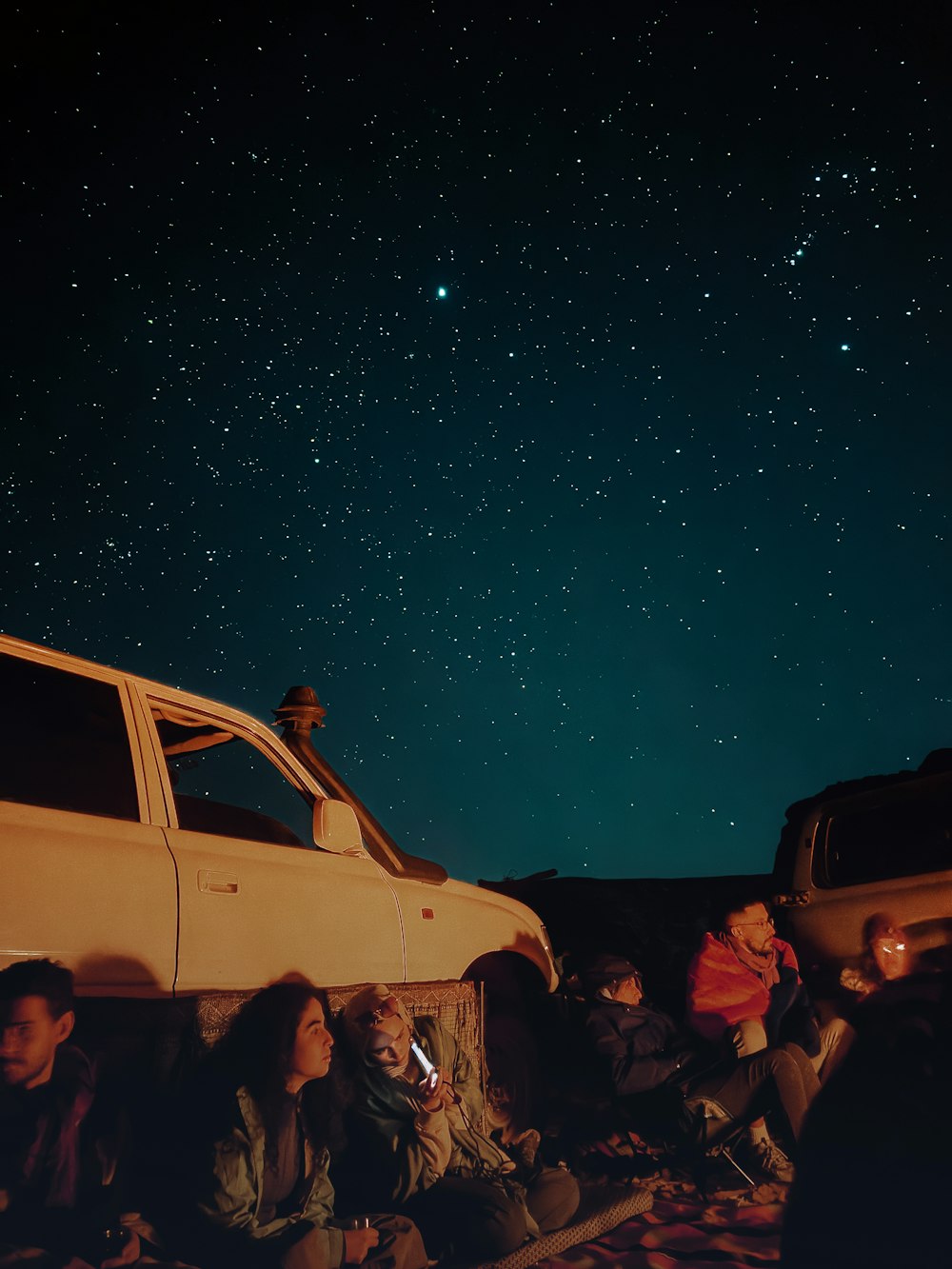 a group of people sitting next to a car under a night sky