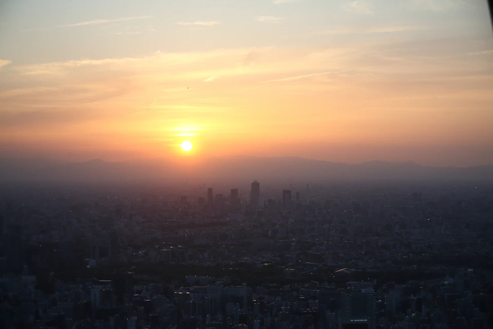 The image depicts a cityscape at sunset, with the sun setting behind the horizon and casting a warm glow over the urban landscape. The sky is painted in shades of orange and yellow, creating a stunning backdrop for the city's buildings. The perspective from above provides an expansive view of the city below, showcasing its architectural diversity and scale.