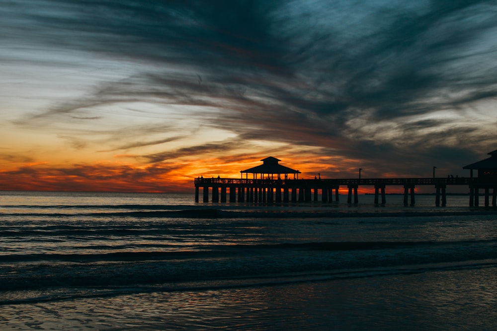 the sun is setting over the ocean with a pier in the distance