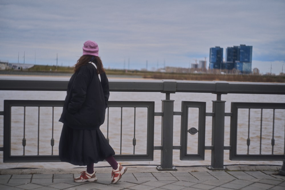 a woman in a black coat and pink hat is walking by the water
