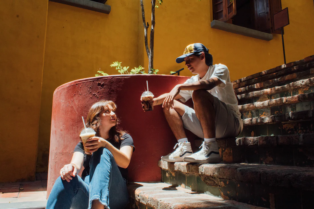 Deux jeunes adultes assis sur des marches devant un mur ocre, une boisson à la main, en pleine conversation détendue.