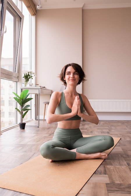 Woman in a green top sitting with hands in prayer on a mat in a bright home living room