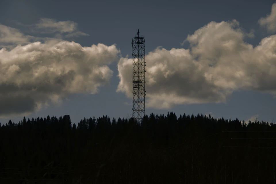 The image depicts a tall tower with multiple levels, standing against a backdrop of a cloudy sky and lush green trees. The tower is constructed from metal and features a weather vane on top. The perspective of the photo is taken from below, looking up at the tower, which gives it an imposing presence in the landscape.