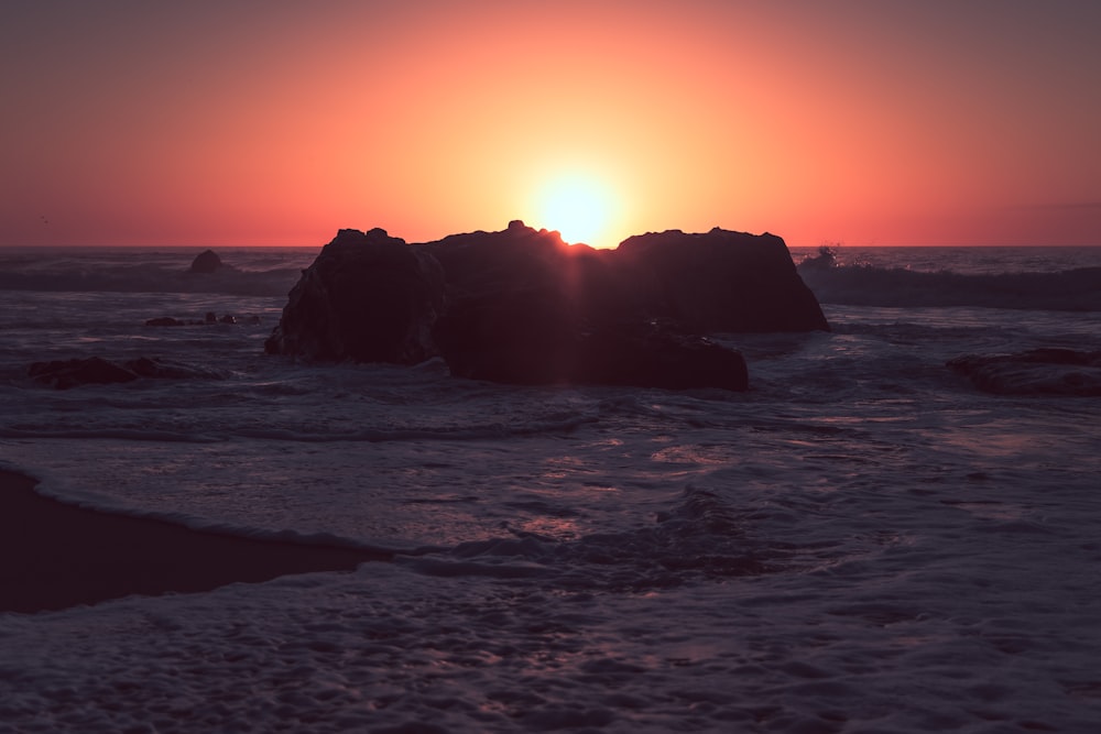 the sun is setting over the ocean with rocks in the foreground