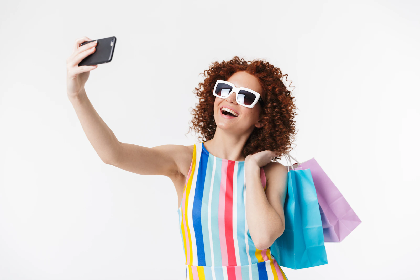 A smiling woman in sunglasses taking a selfie while holding shopping bags