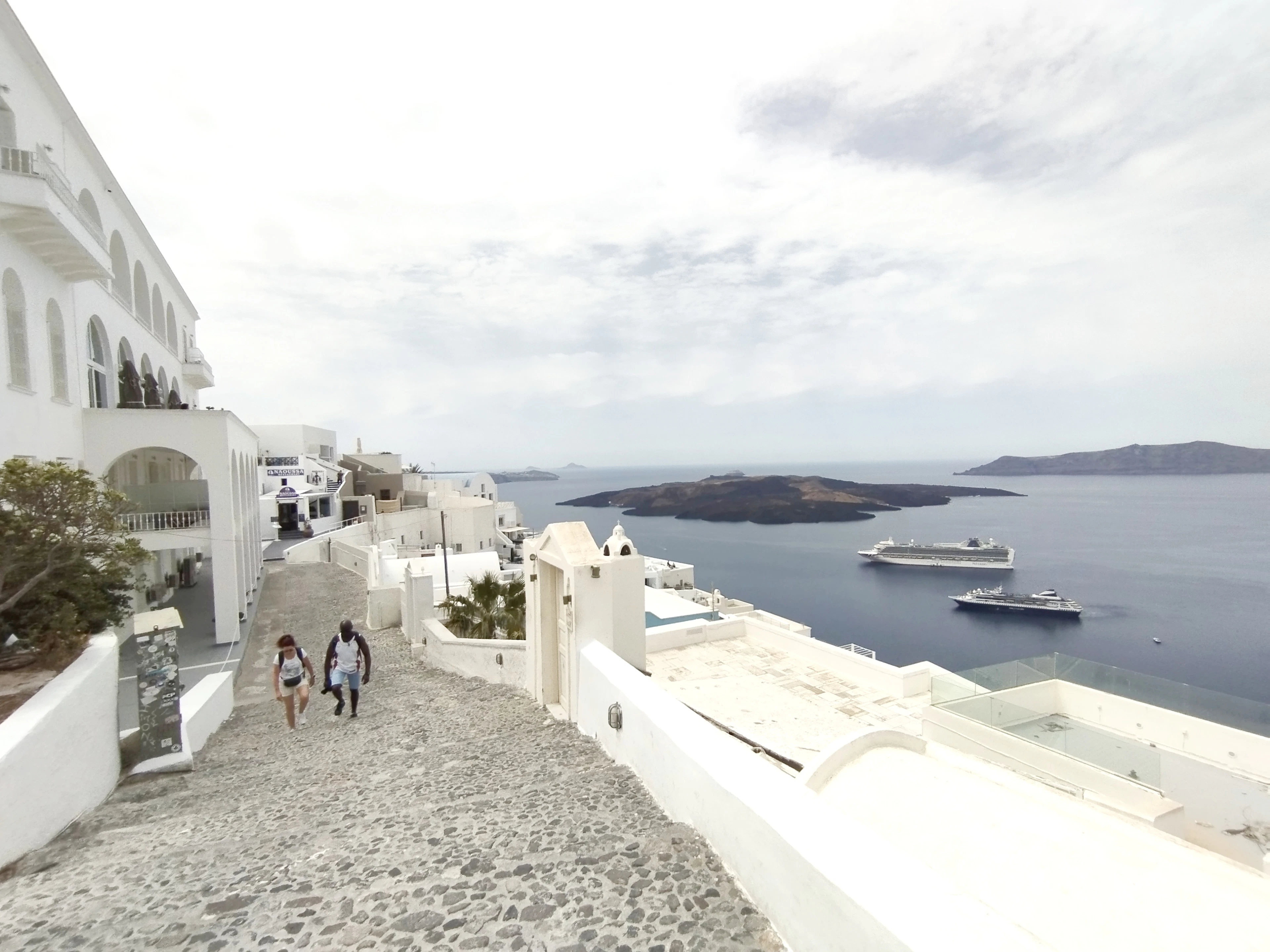Walkers on the cobbled clifftop path through Fira, Santorini, passing whitewashed terraces with cruise ships anchored in the caldera below