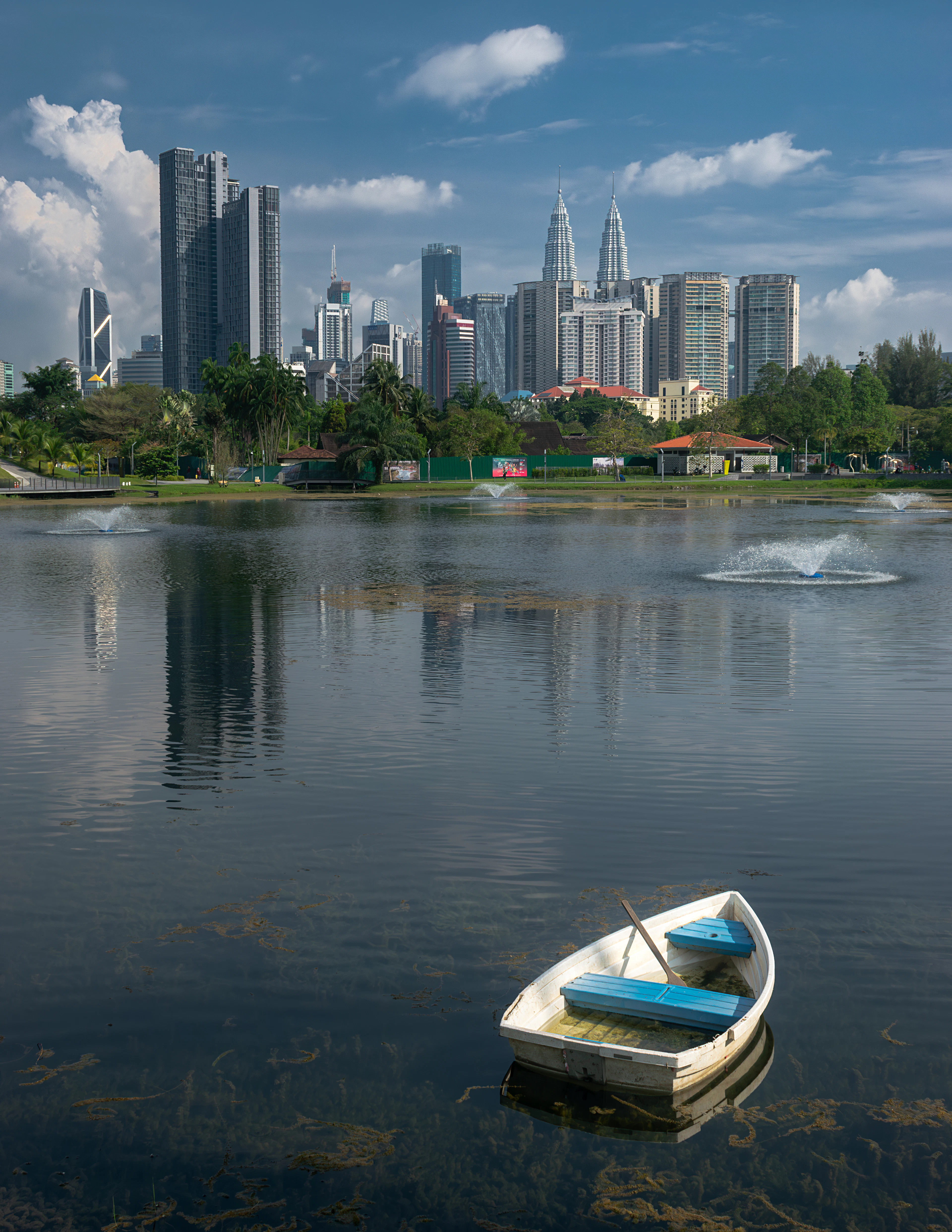 A small white rowing boat drifting on the still water of Titiwangsa Lake in Kuala Lumpur, park fountains playing across the middle distance and the city skyline rising beyond the treeline under piled white cloud.