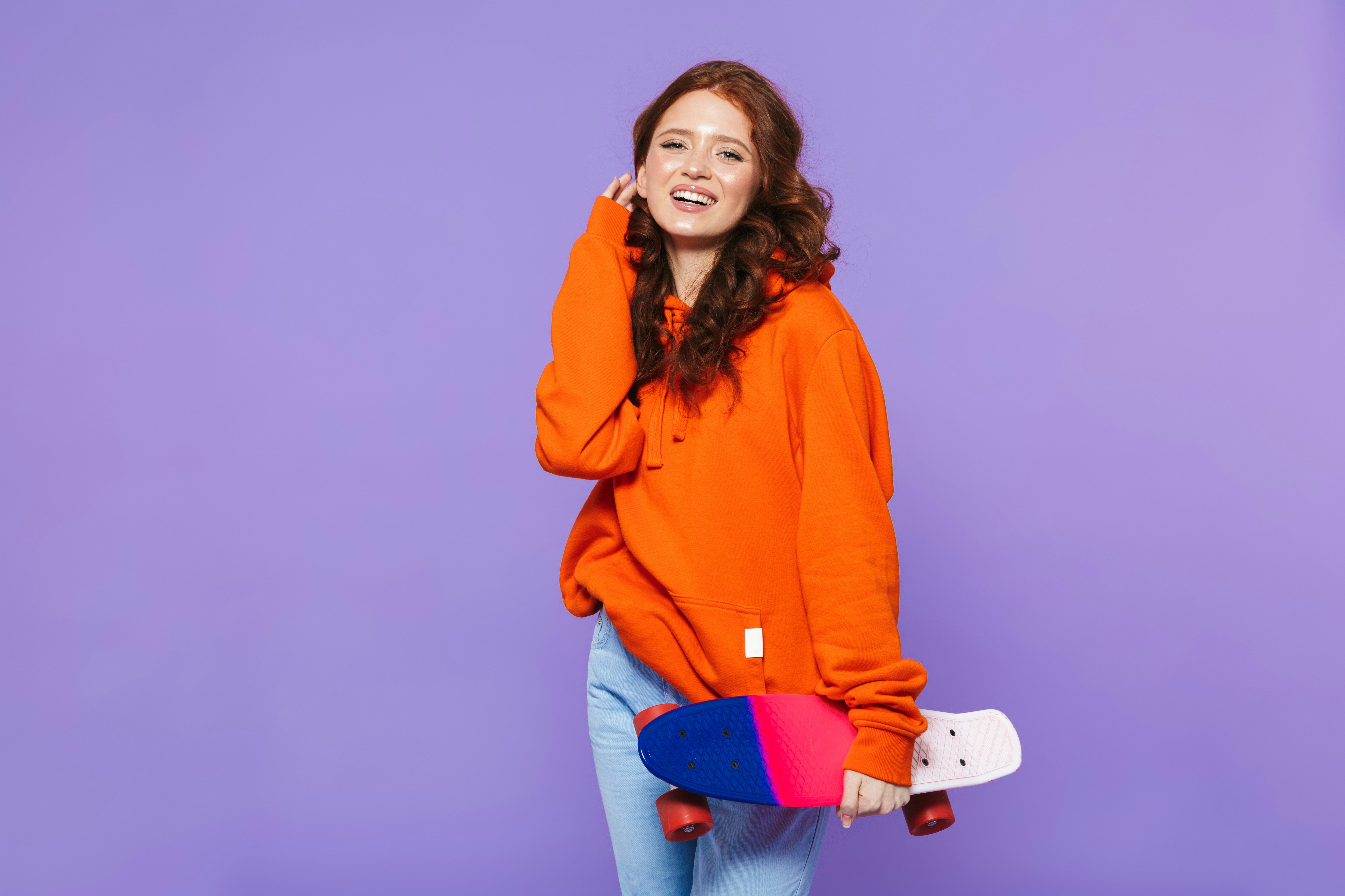 A smiling woman in an orange sweater against a lilac wall