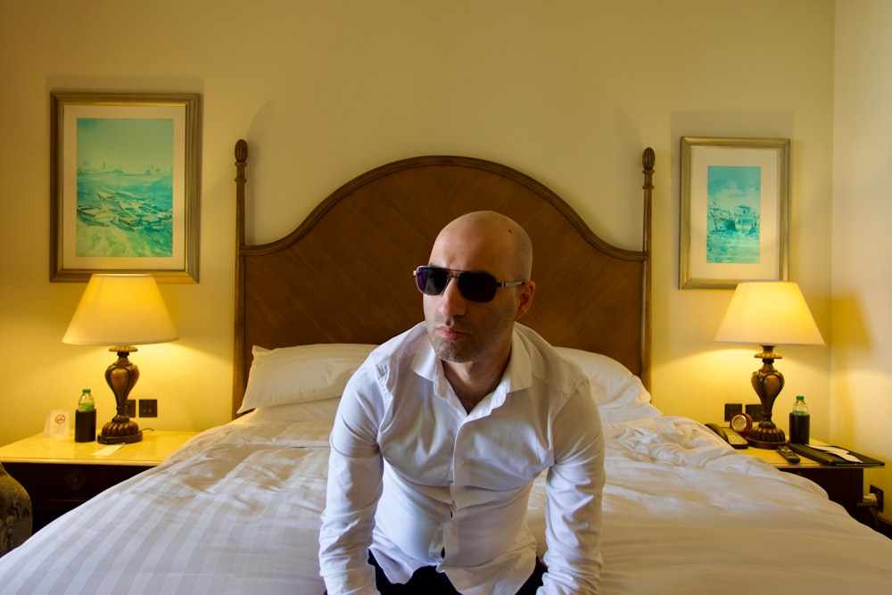 a bald man in sunglasses sitting on a bed