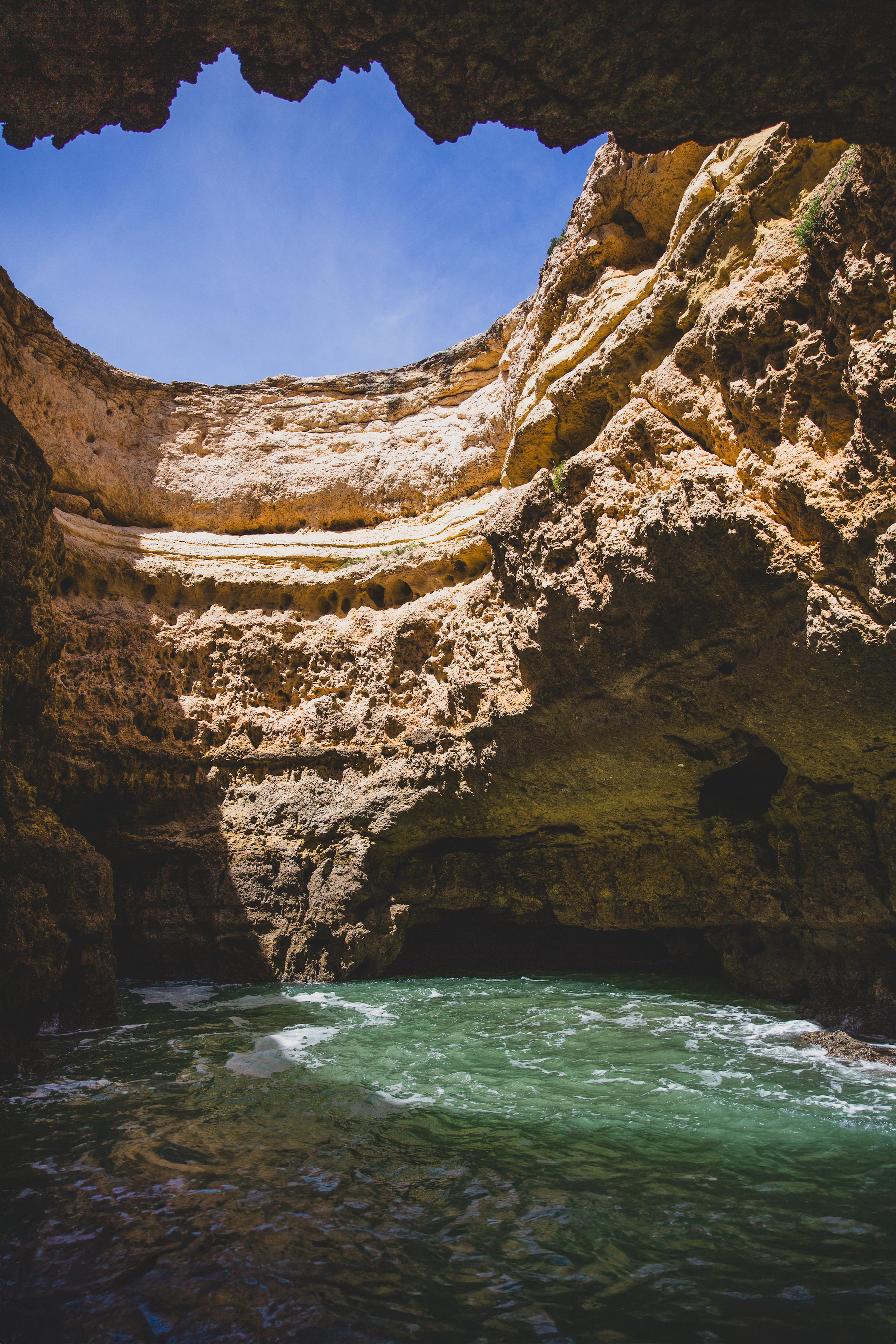 Inside the Benagil sea cave in the Algarve, looking up through the circular opening in the sandstone roof to blue sky, with green-tinged water washing the cave floor