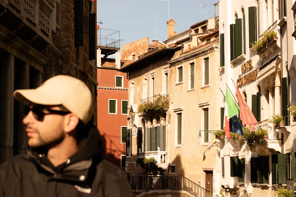 a man wearing a hat and sunglasses standing in front of a row of buildings