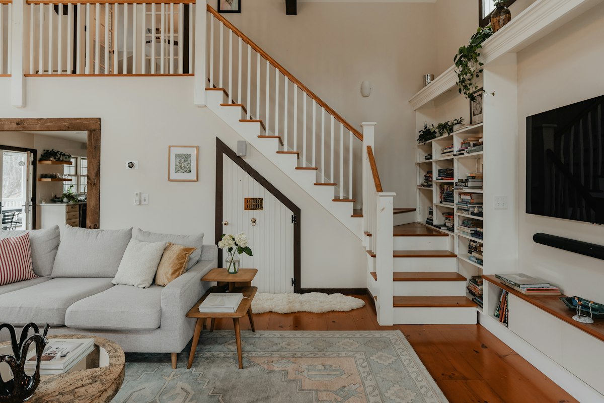Living con sofá gris y escalera blanca de peldaños de madera; debajo de la escalera hay una puerta de placard con forma de triángulo y al costado una biblioteca empotrada llena de libros