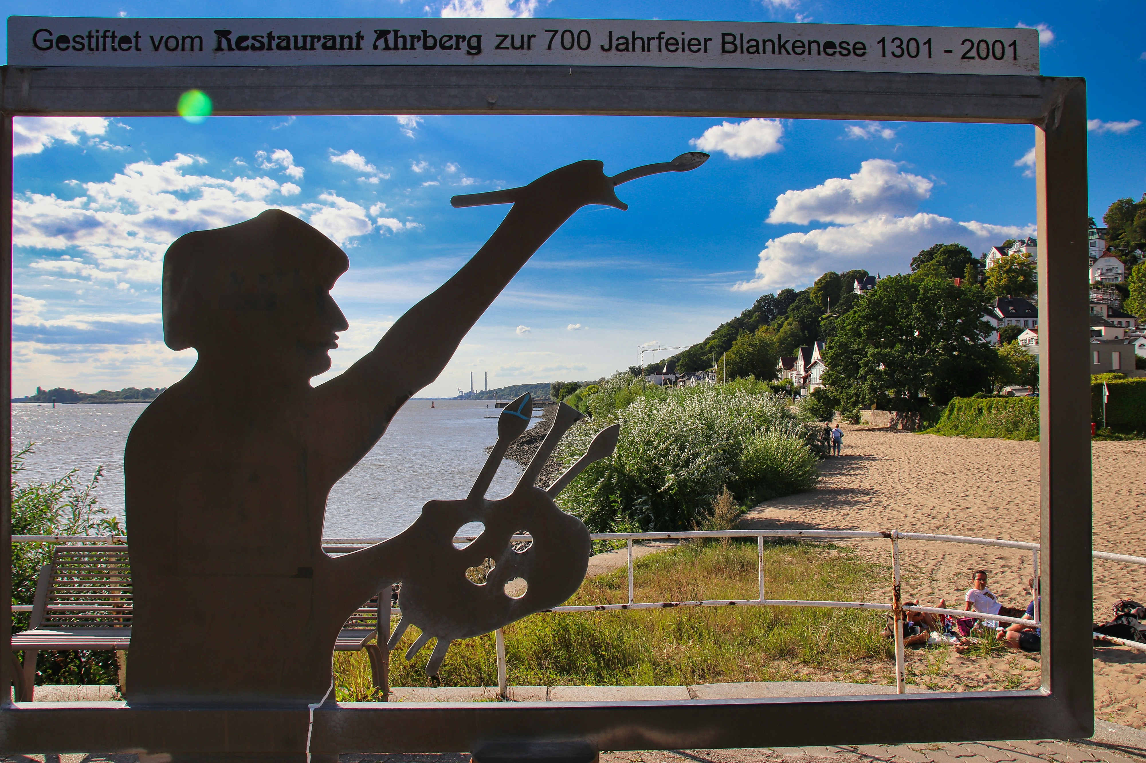 Metal artist sculpture overlooking a river, sandy beach, and hillside.