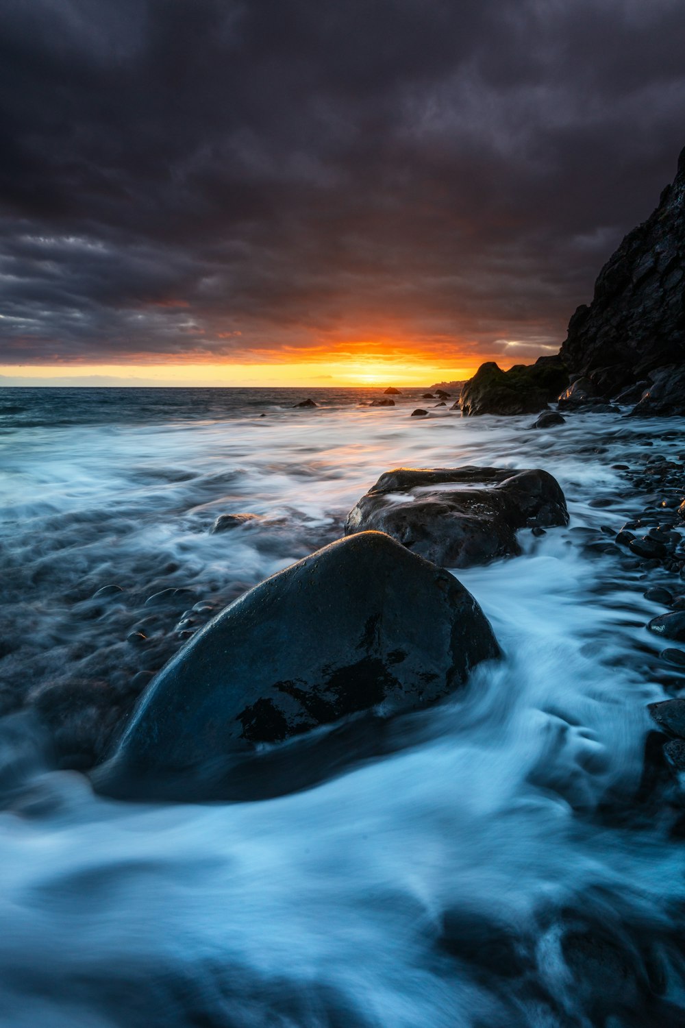 the sun is setting over the ocean with rocks in the foreground