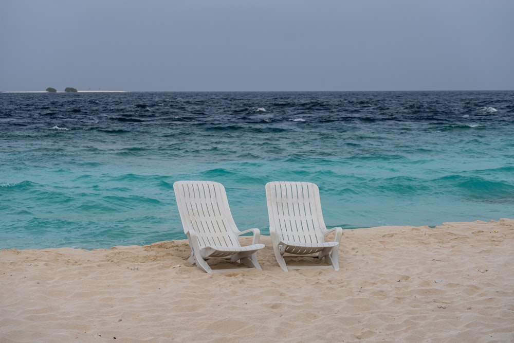 two white chairs sitting on top of a sandy beach