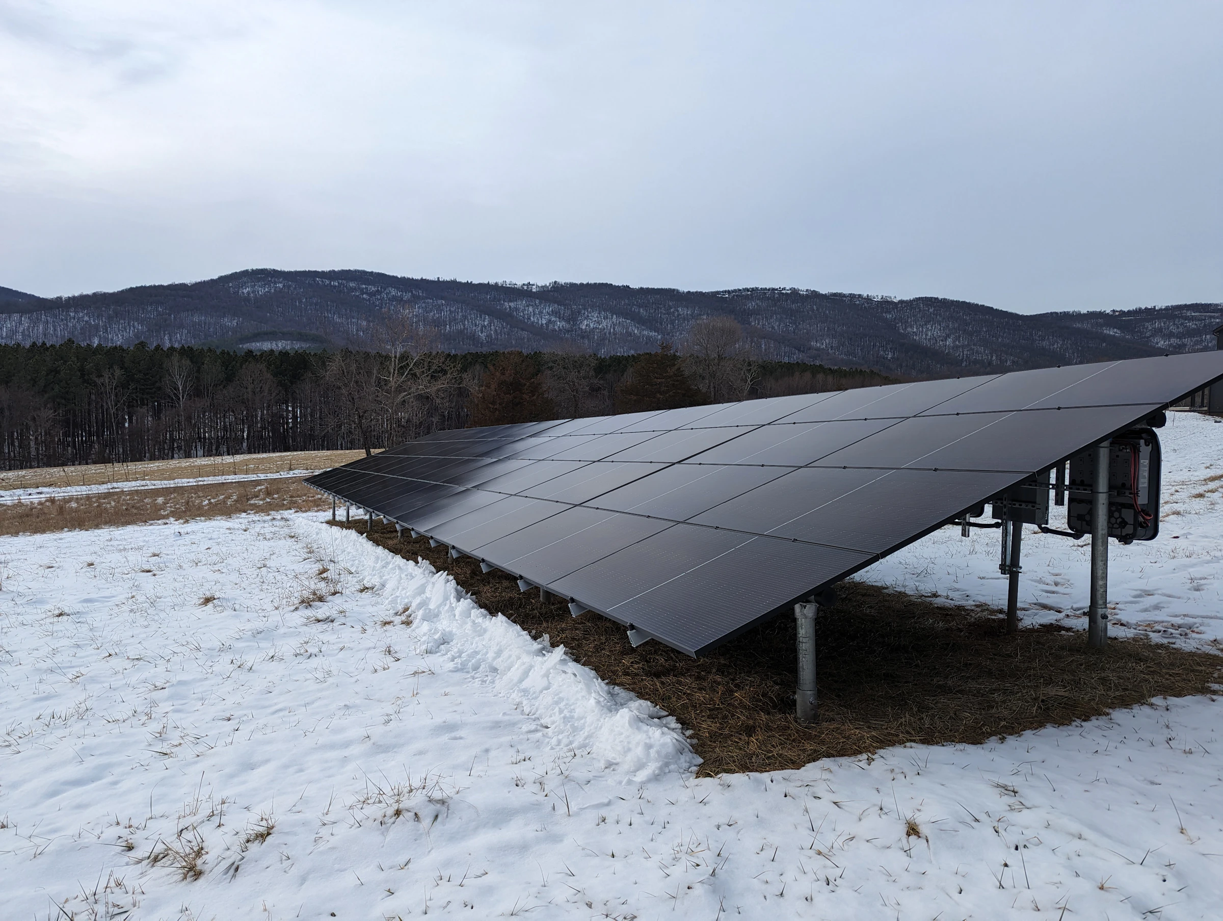 Ground-mounted solar array in a snowy field below forested hills