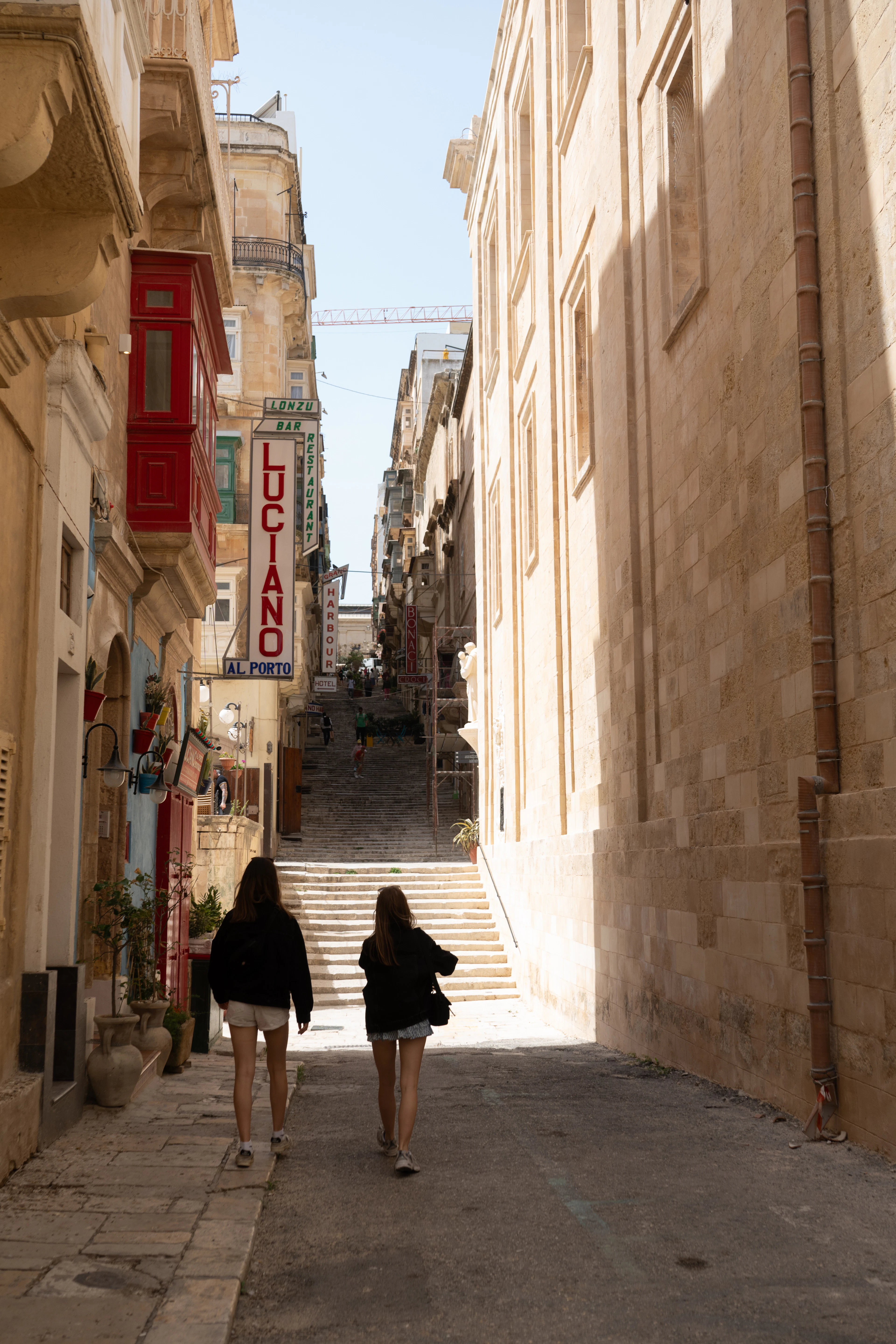 Two women walk down a near-empty Valletta side street towards the long flight of limestone steps that drops to the harbour, a red timber balcony and the vertical sign of a small bar-restaurant overhead.