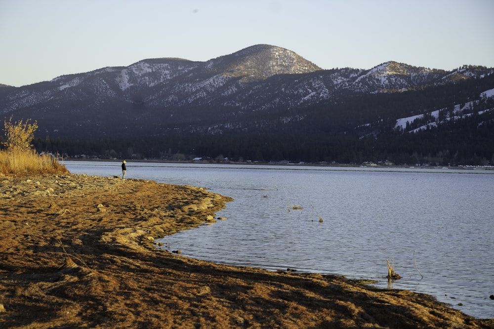 a man standing on the shore of a lake with mountains in the background