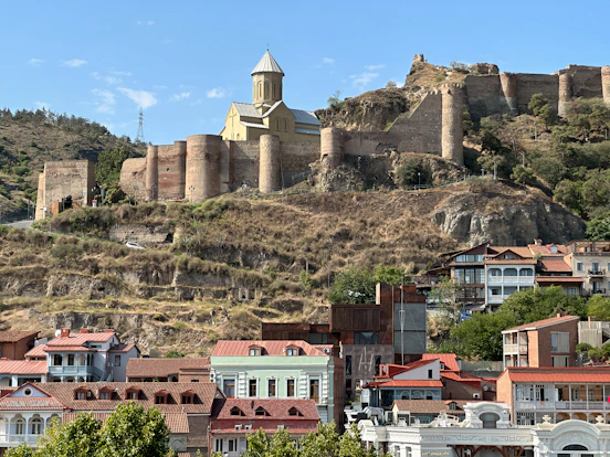  A stone fortress sits on a grassy hill above colorful buildings in the city of Tbilisi Georgia.