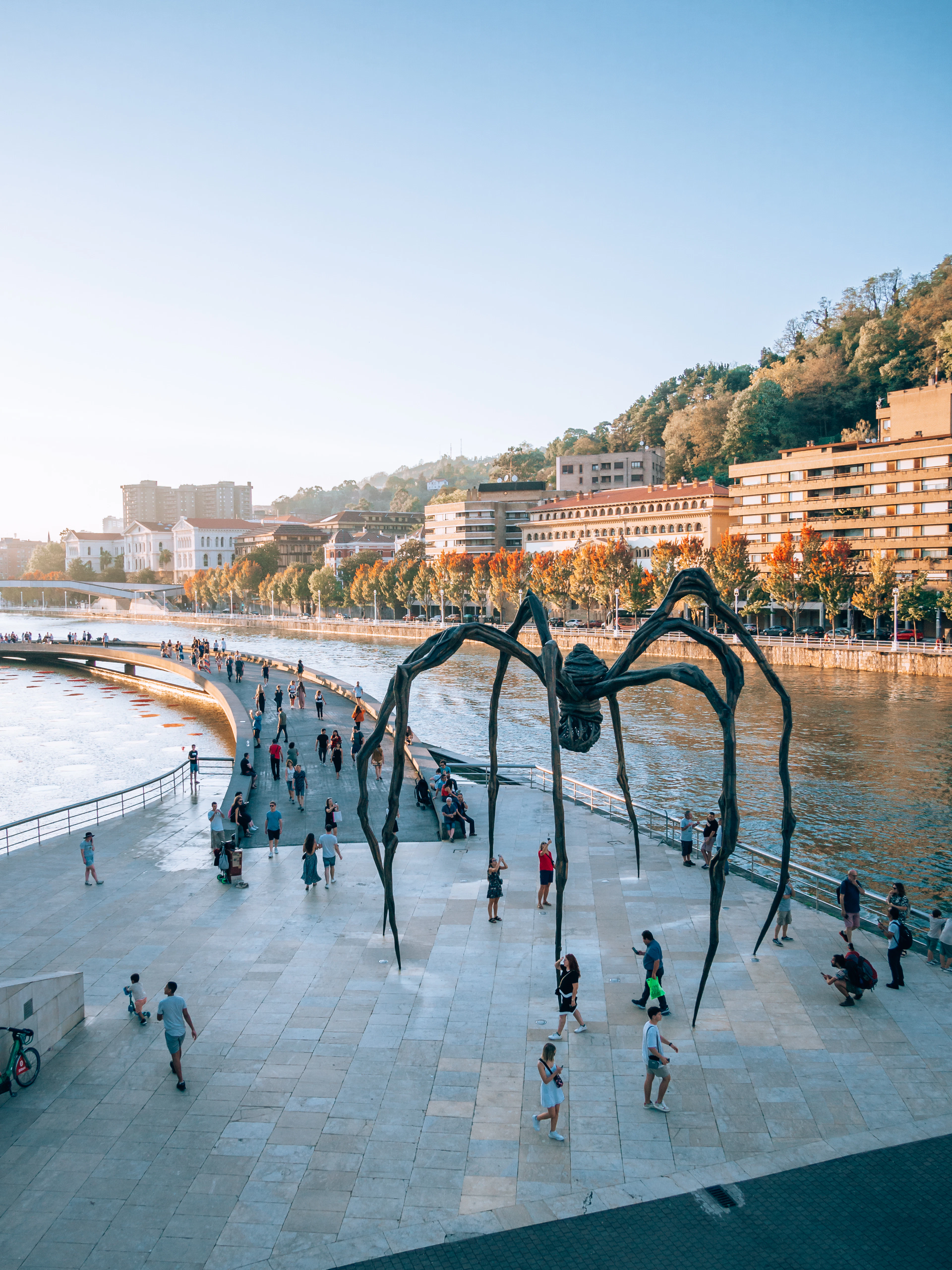 Visitors on the riverside terrace beside the Guggenheim Bilbao, walking beneath a monumental bronze spider sculpture