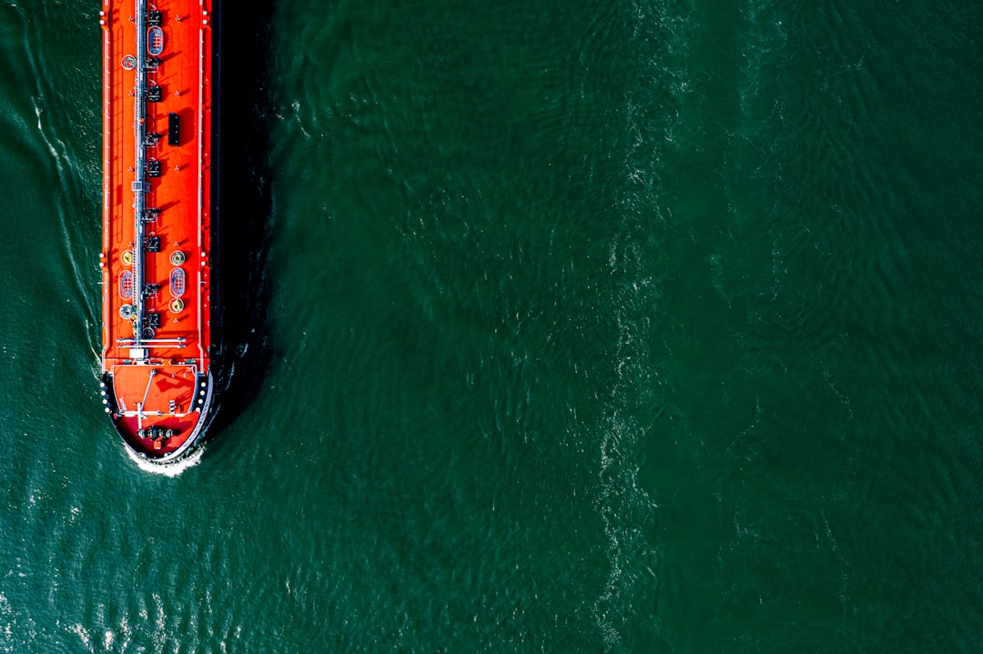 From an aerial perspective, a vivid red cargo ship cuts through the green waters, its wake creating a stark contrast. This image encapsulates the vitality of maritime logistics and the movement of goods along crucial waterways.