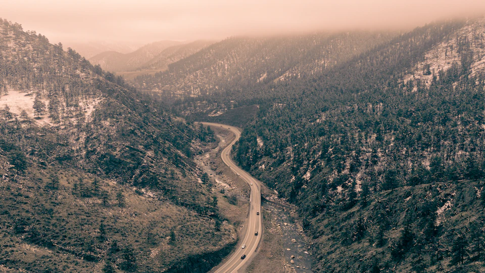 The image depicts a winding road cutting through a mountainous landscape, with trees and foggy mountains on either side. The road is flanked by lush green trees that add depth to the scene. The perspective suggests an aerial view from above, providing a comprehensive look at the winding path of the mountain road.