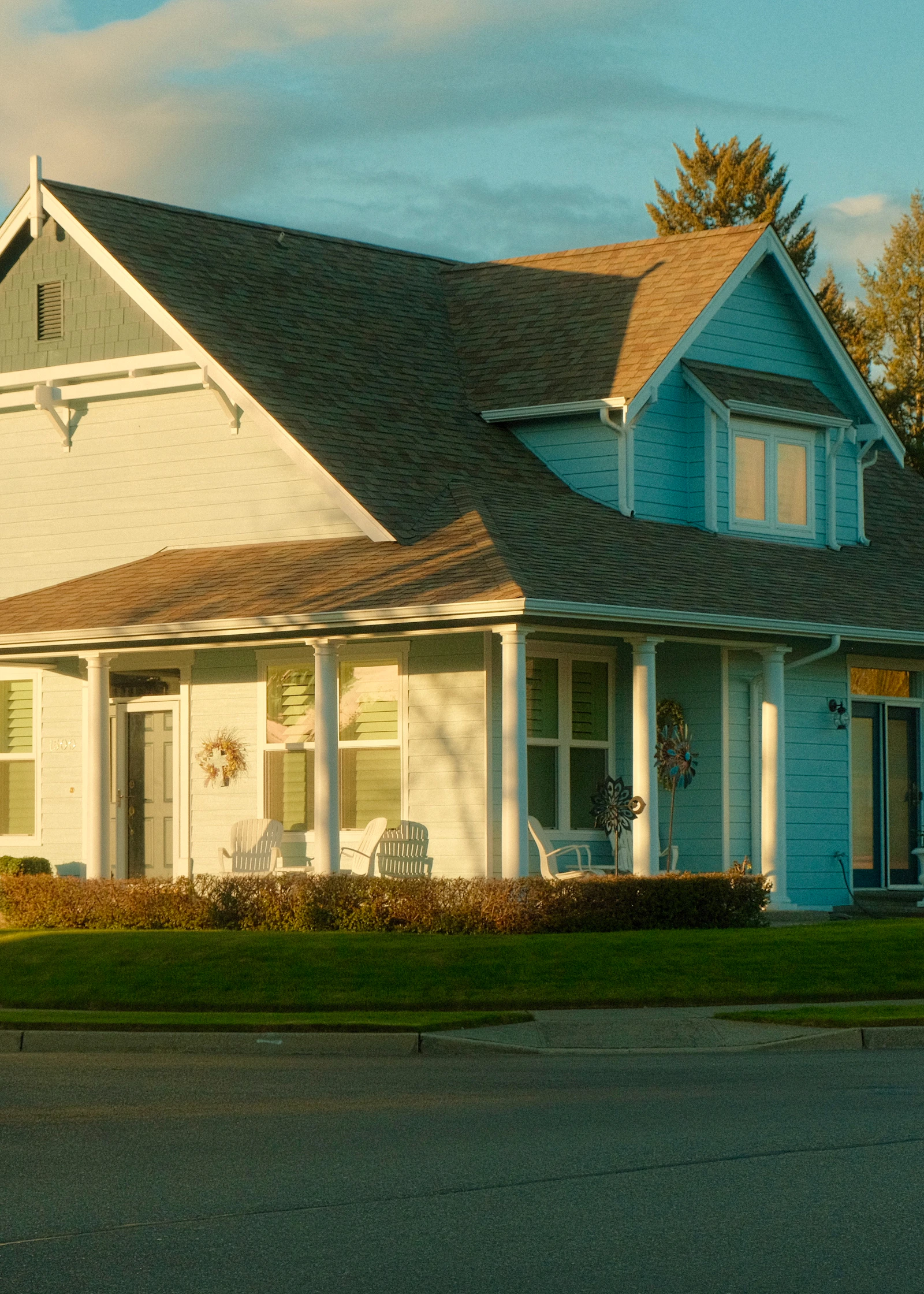 Light-blue two-story house with a wraparound porch and dormer windows catching warm golden-hour light