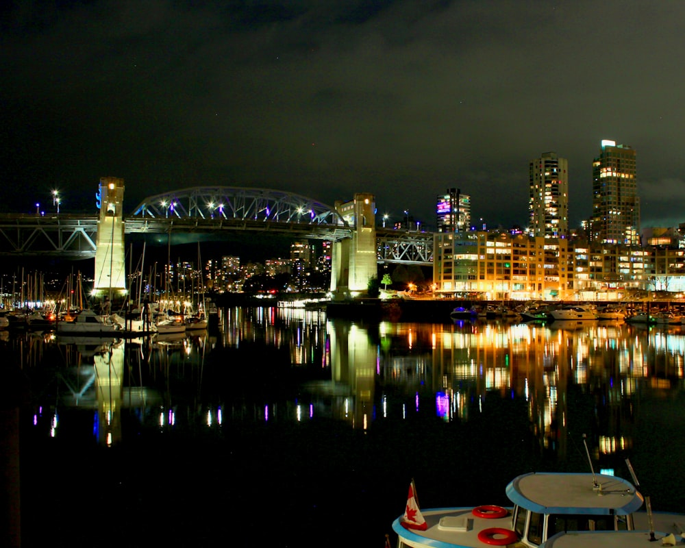 a night scene of a city with a bridge and boats in the water
