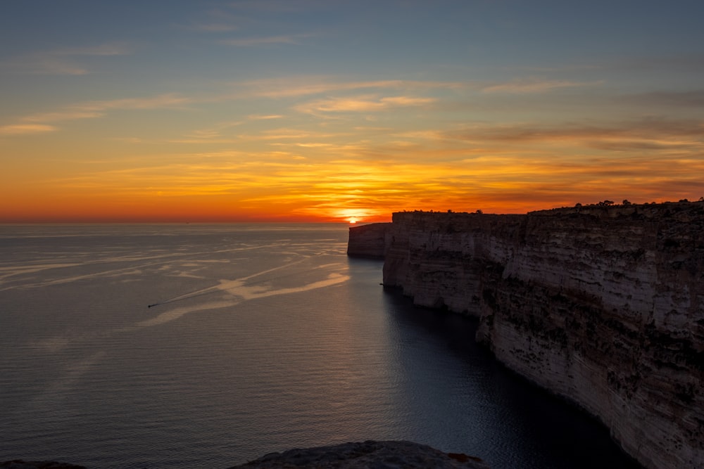 the sun is setting over the water at the edge of a cliff