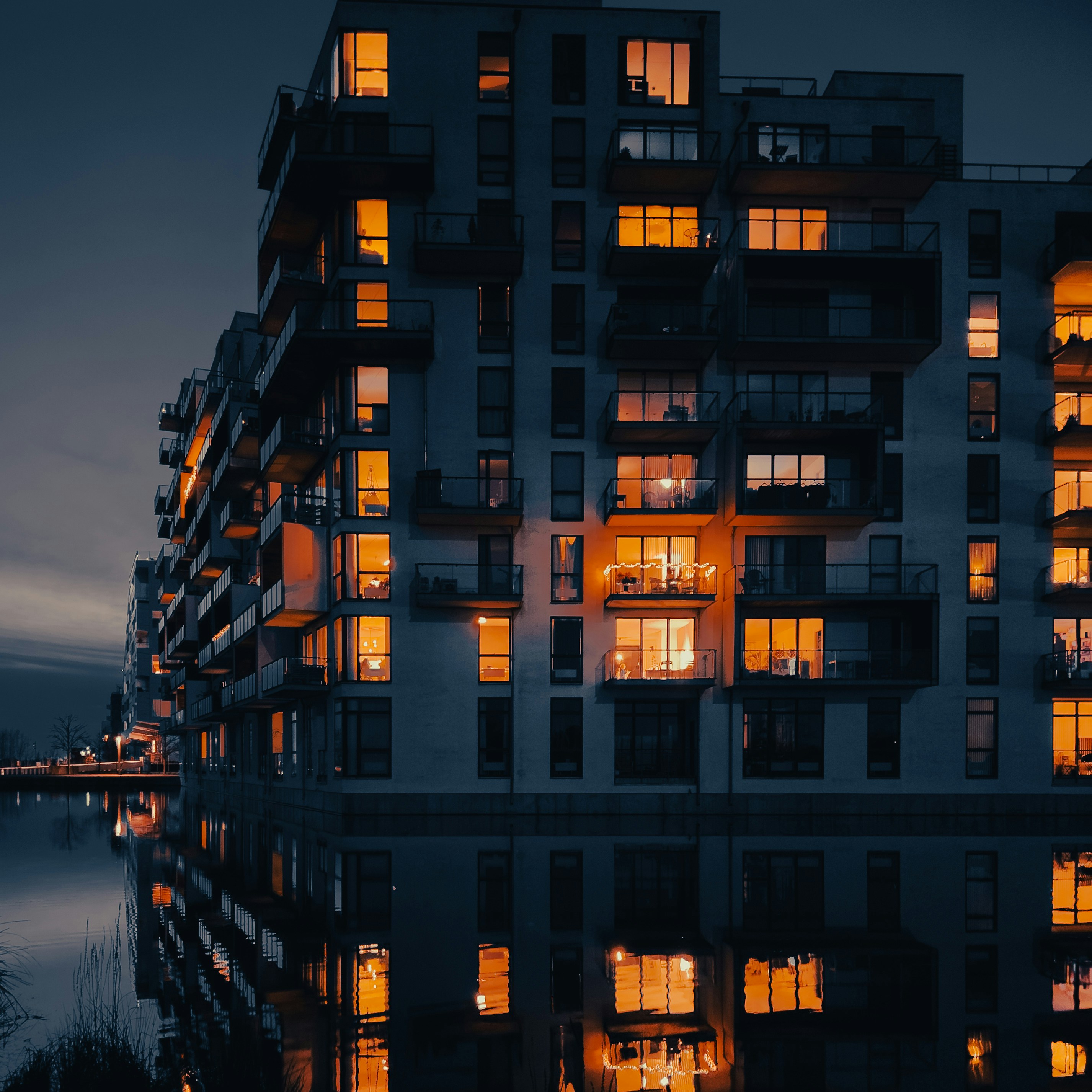 Waterfront luxury apartment balcony overlooking marina at dusk, dark water, warm golden lights reflecting, moody atmospheric haze