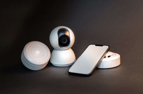 A smartphone next to a wireless home security camera on a white surface