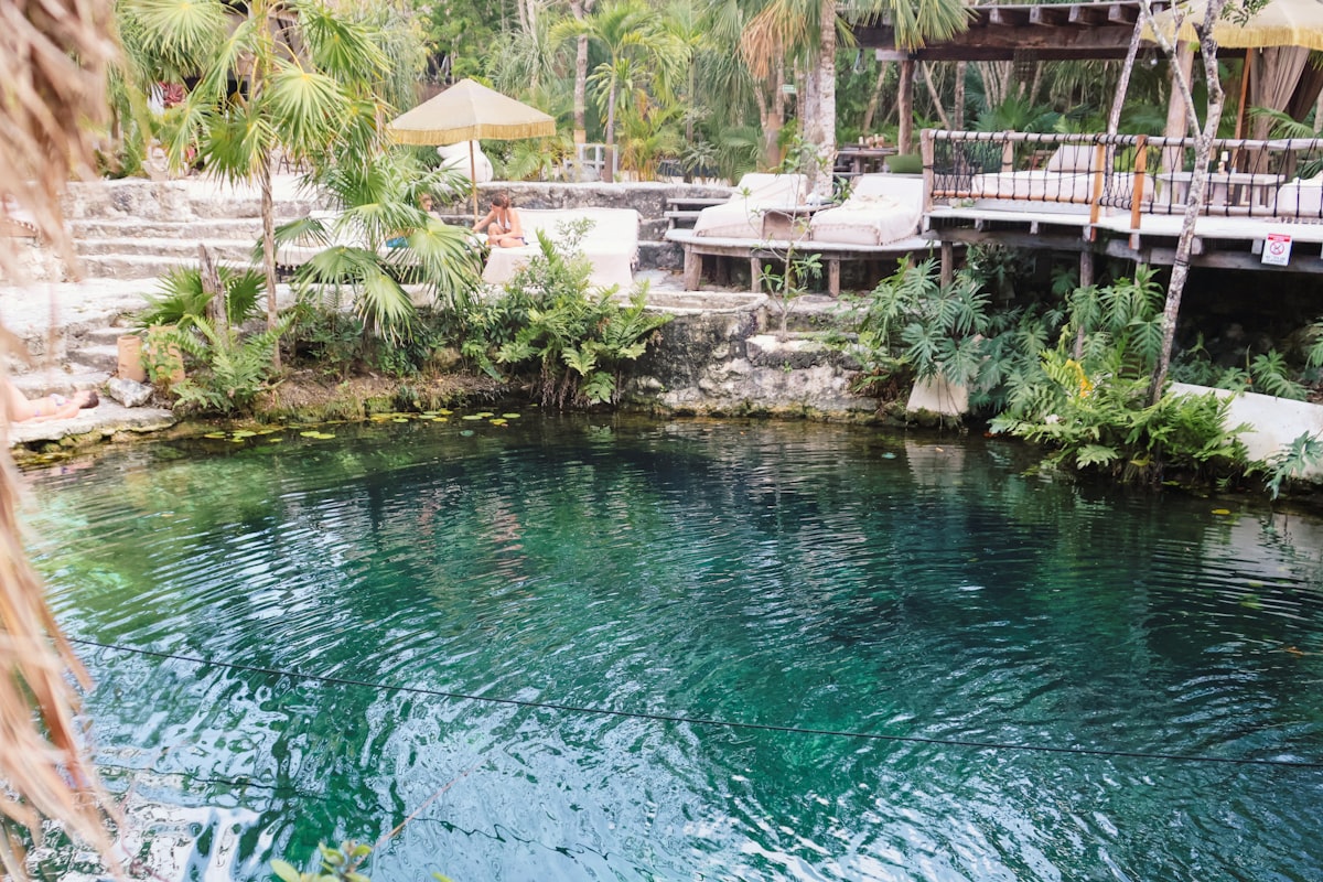Tulum October — a lush cenote sanctuary with turquoise water surrounded by tropical trees and umbrellas as the wet season winds down