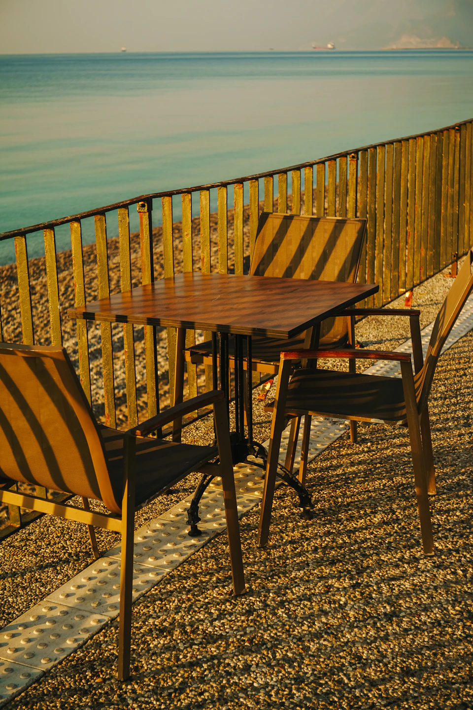 Two wooden tables with chairs on pebbled ground near the sea's edge