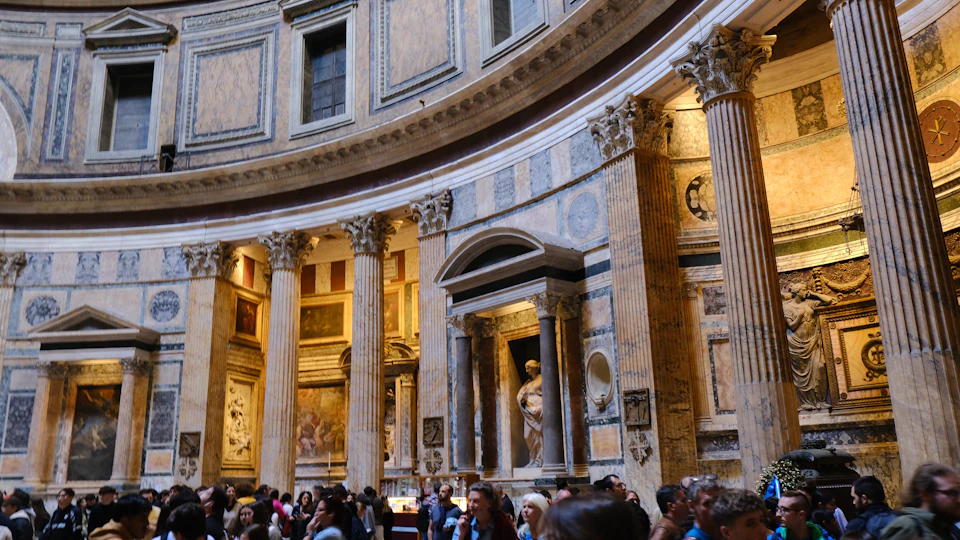 The image depicts a large, ornate room with a curved ceiling and walls adorned with intricate designs. The room is filled with people, creating a sense of scale and grandeur. The perspective of the photo is from below, looking up at the towering columns that support the roof. The predominant colors in the scene are beige and brown, reflecting the natural hues found in stone and marble.
