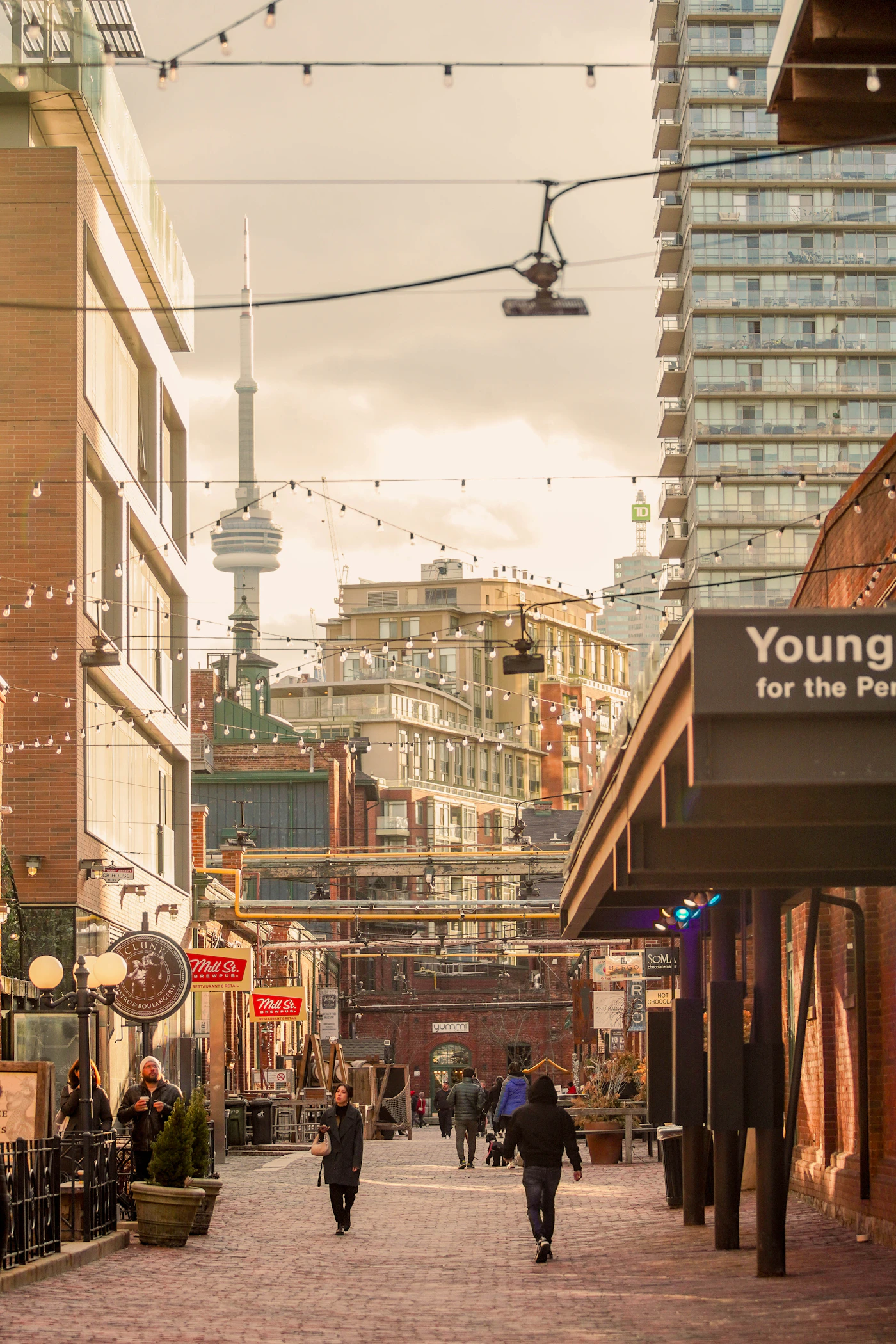 Toronto's heritage entertainment district with Victorian brick buildings, overhead string lights, and the CN Tower visible above the roofline in the distance