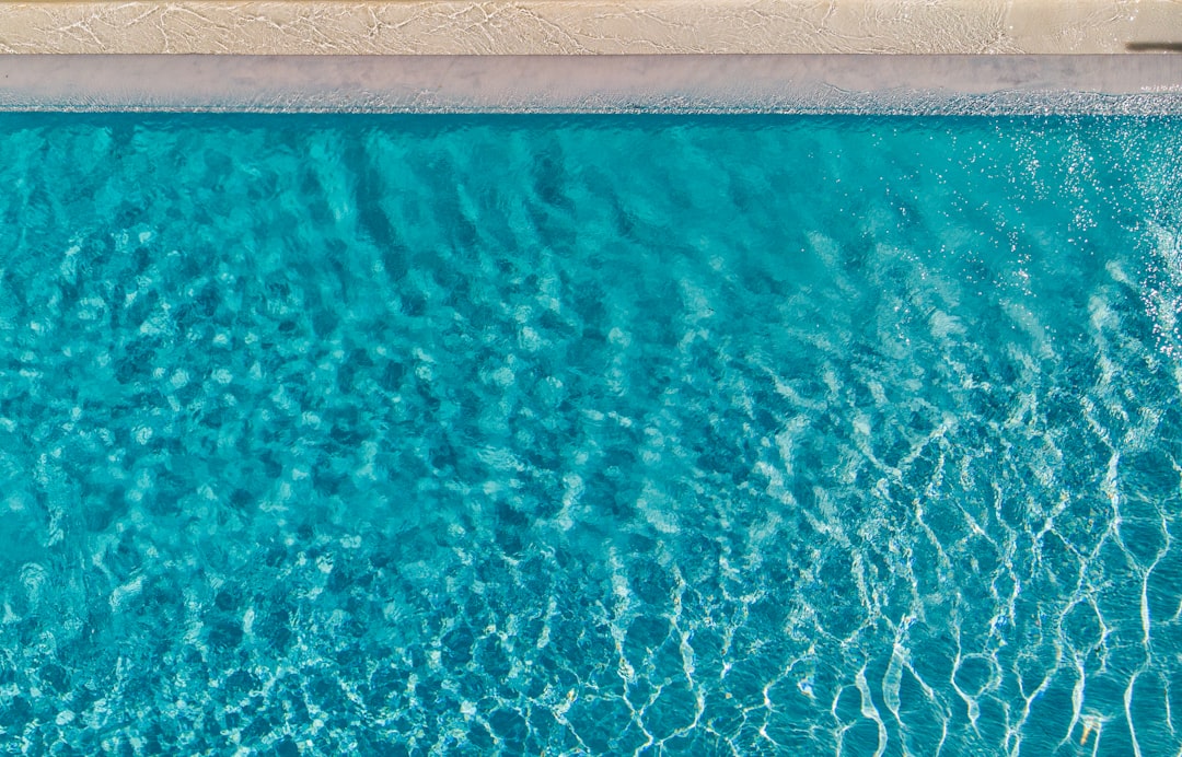 An aerial view of a swimming pool with crystal clear turquoise water. Sunlight dances across the surface, creating a pattern of shimmering ripples. The pool's edge is visible, delineating the refreshing water from the surrounding area.