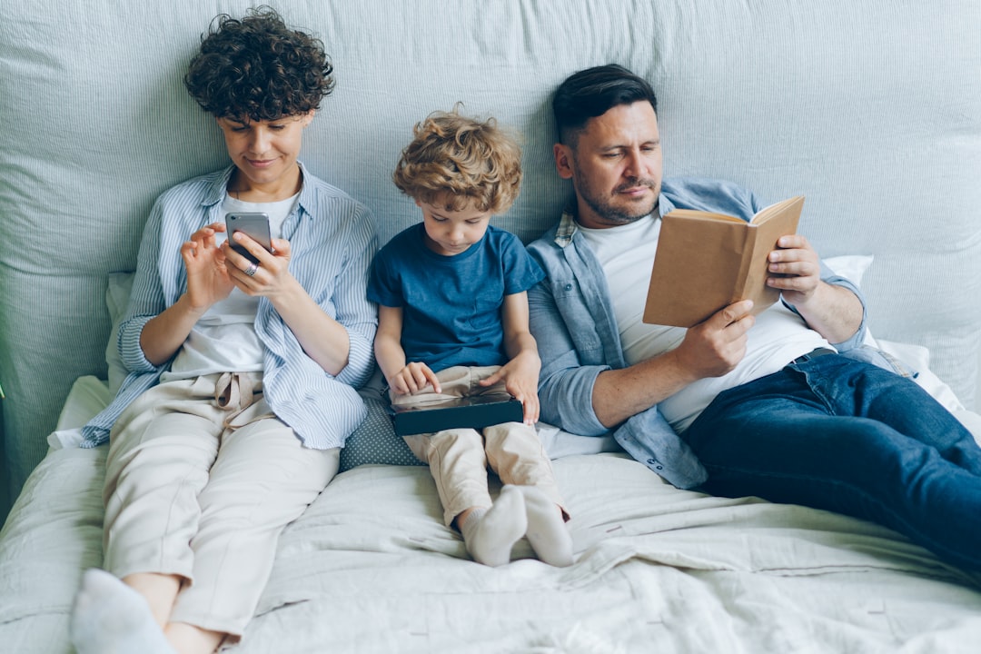 a man and two children are sitting on a bed, Mother using smartphone son holding tablet father reading book in bed at ho