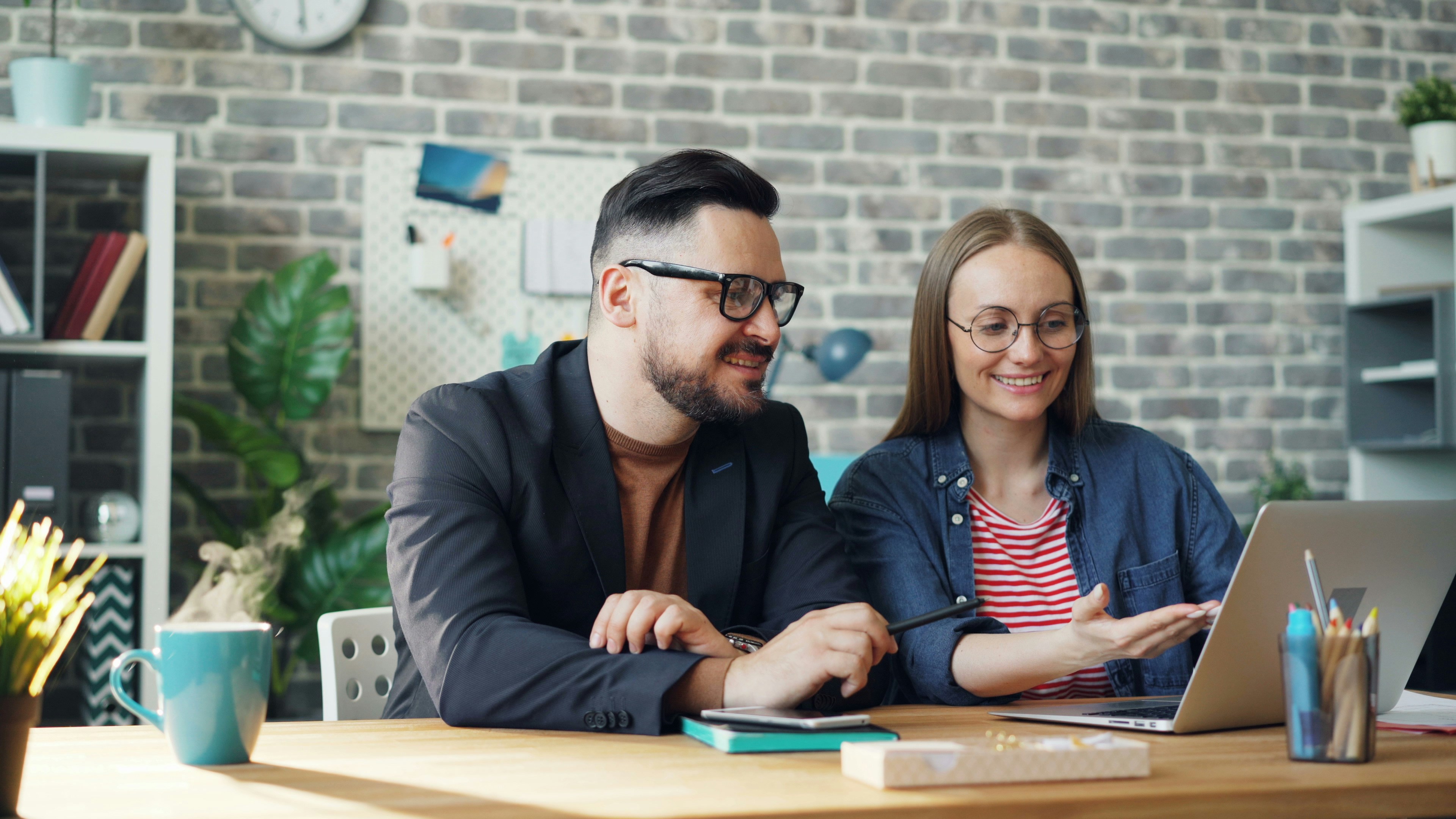 Male and female coworkers are discussing work sitting at desk with laptop together talking smiling. Cooperation, business people and communication concept.